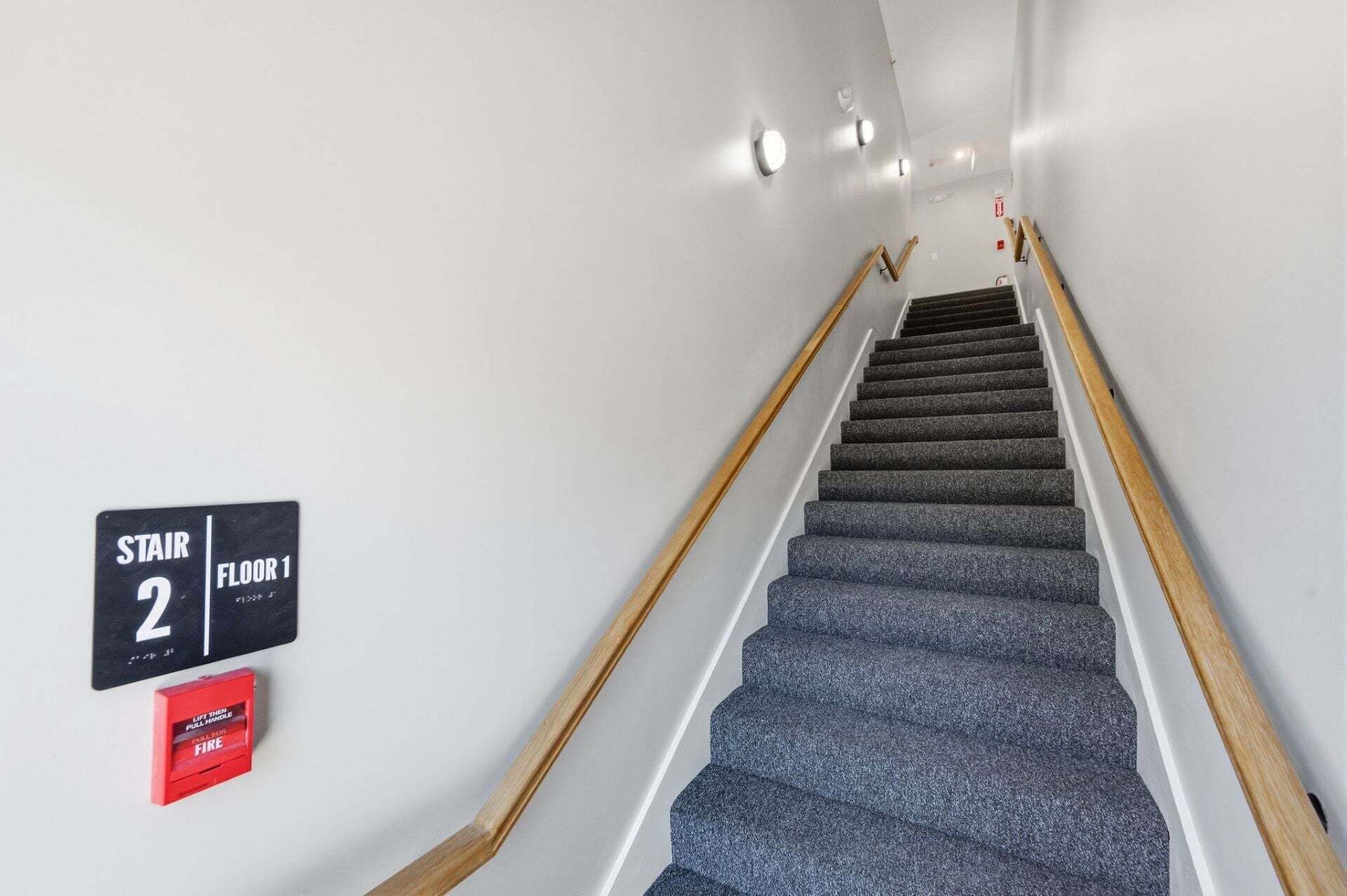A staircase leading to the first floor with a handrail on both sides and a fire pull handle on the wall.