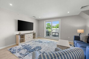 A living room with a TV, a sofa, a coffee table, and a view through a sliding door. - Charming Cape Apartment Walk to Beach & Town (Harwich Port)