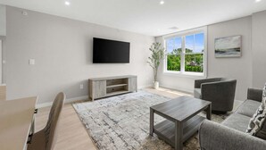 Een moderne woonkamer met een flatscreen televisie, grijze banken, een salontafel en een groot raam met uitzicht.