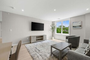 A modern living room with a flat-screen TV, grey sofas, a coffee table, and a large window with a view. - Relaxing Harwich Port Apartment Near Beach & Downtown (Harwich Port)