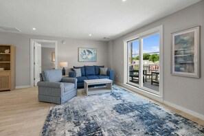 A spacious living room with a blue sofa, striped armchair, and a coffee table. There is a large window providing a view of the outdoors. - Relaxing Harwich Port Apartment Near Beach & Downtown (Harwich Port)