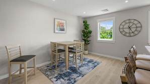 Een eetgedeelte met een tafel en stoelen, een plant in pot, een klok en een raam met uitzicht.