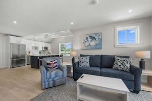 A spacious living area with a sofa, armchair, and coffee table, a kitchen with modern appliances, and a large window providing natural light. - Beachside Harwich Port Apartment Near Town (Harwich Port)