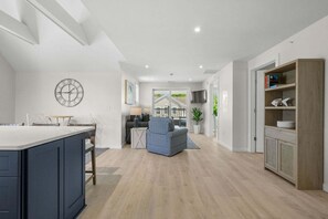 A modern kitchen with a central island, a dining area with a table and chairs, a living room with a sofa and a TV, and a wooden cabinet with shelves. - Beachside Harwich Port Apartment Near Town (Harwich Port)