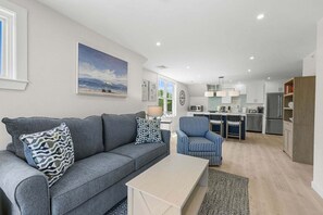 Ein modernes Wohnzimmer mit einem grauen Sofa, gestreiften Sesseln und einem Couchtisch. Im Hintergrund ist eine Küche mit weißen Schränken und Edelstahlgeräten erkennbar.