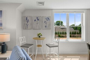 A room with a white ceiling, a window with a view of a house and trees, a round table with a vase of flowers, two white chairs, and a blue-striped sofa. - Harwich Port Apartment Just Minutes to Beach & Town (Harwich Port)