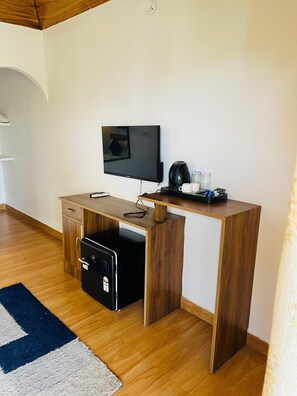 Una habitación compacta con escritorio de madera, televisor, cafetera y un refrigerador pequeño.