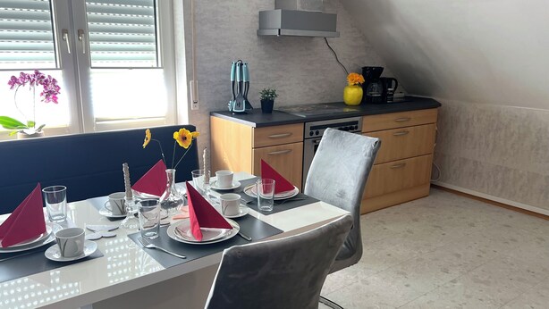 Ein Essbereich mit Tisch für zwei Personen, eine Küche im Hintergrund und ein Fenster mit Jalousien.