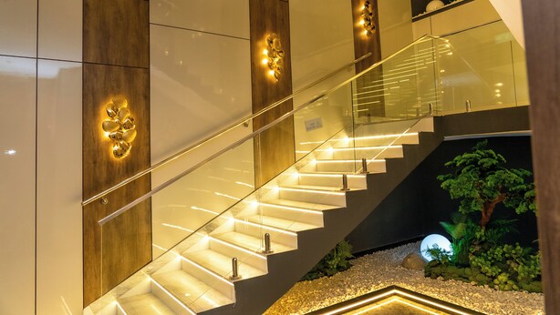Una escalera moderna con barandillas de vidrio y peldaños iluminados, rodeada por una combinación de paneles de madera y blancos.