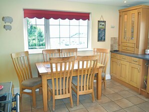 A dining area with a wooden table and chairs, a window with a red valance, and wooden kitchen cabinets. - 3 bedroom accommodation in Braunton (Braunton)