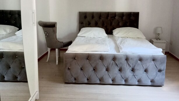 Een slaapkamer met een gestoffeerd hoofdeinde, een grijze fauteuil, een nachtkastje met een lamp, en een grote spiegel aan de muur.