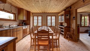 Interior em madeira com uma mesa de jantar posta para seis, armários de madeira e grandes janelas com vista para o exterior.