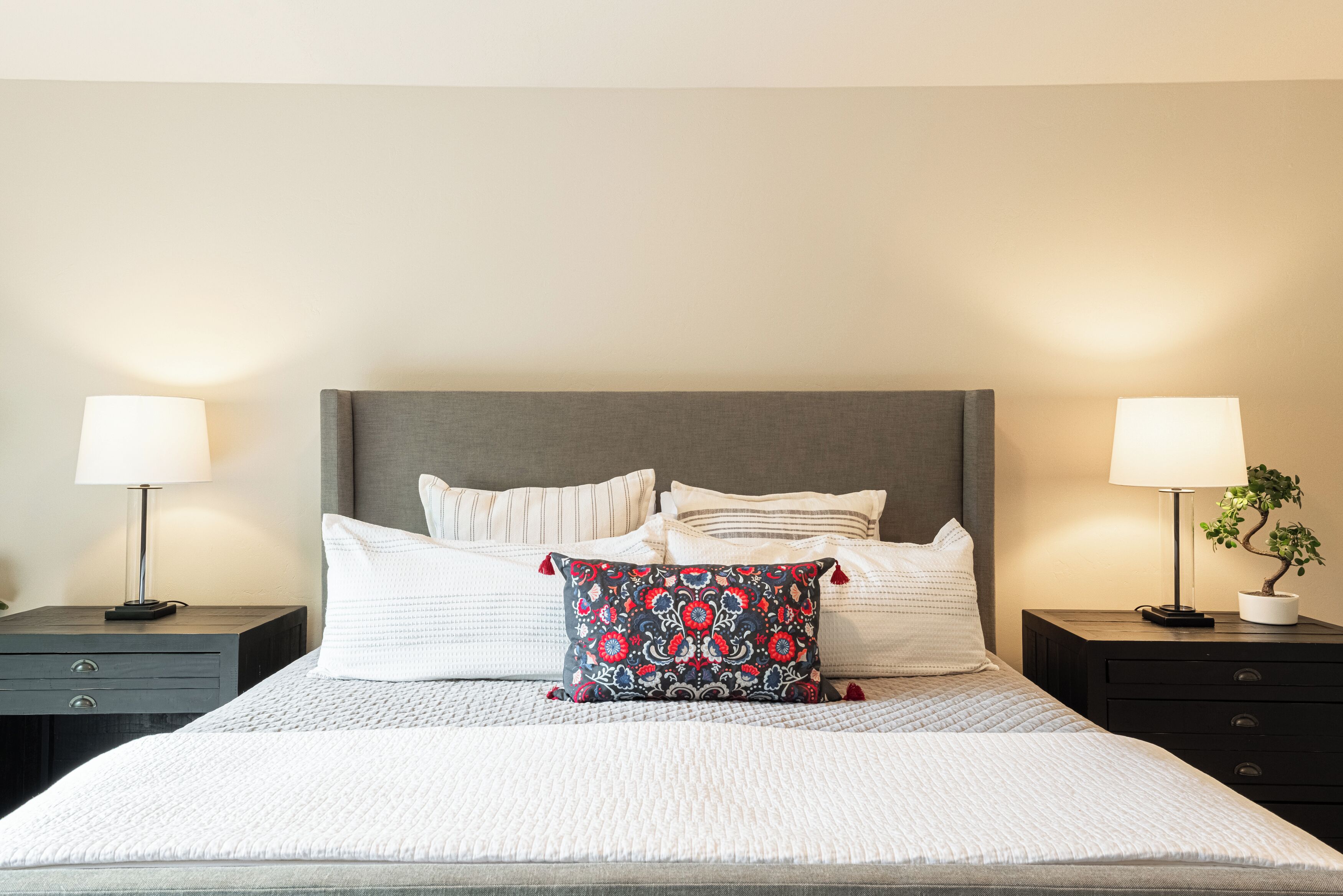 A neatly made bed with a grey headboard, white bedding, and a decorative pillow. There are two bedside tables with lamps, and a small potted plant on one of the tables.