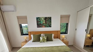Un dormitorio moderno con una cama de madera, dos mesitas de noche con lámparas, un ventanal con vistas a la vegetación y un aire acondicionado de pared.