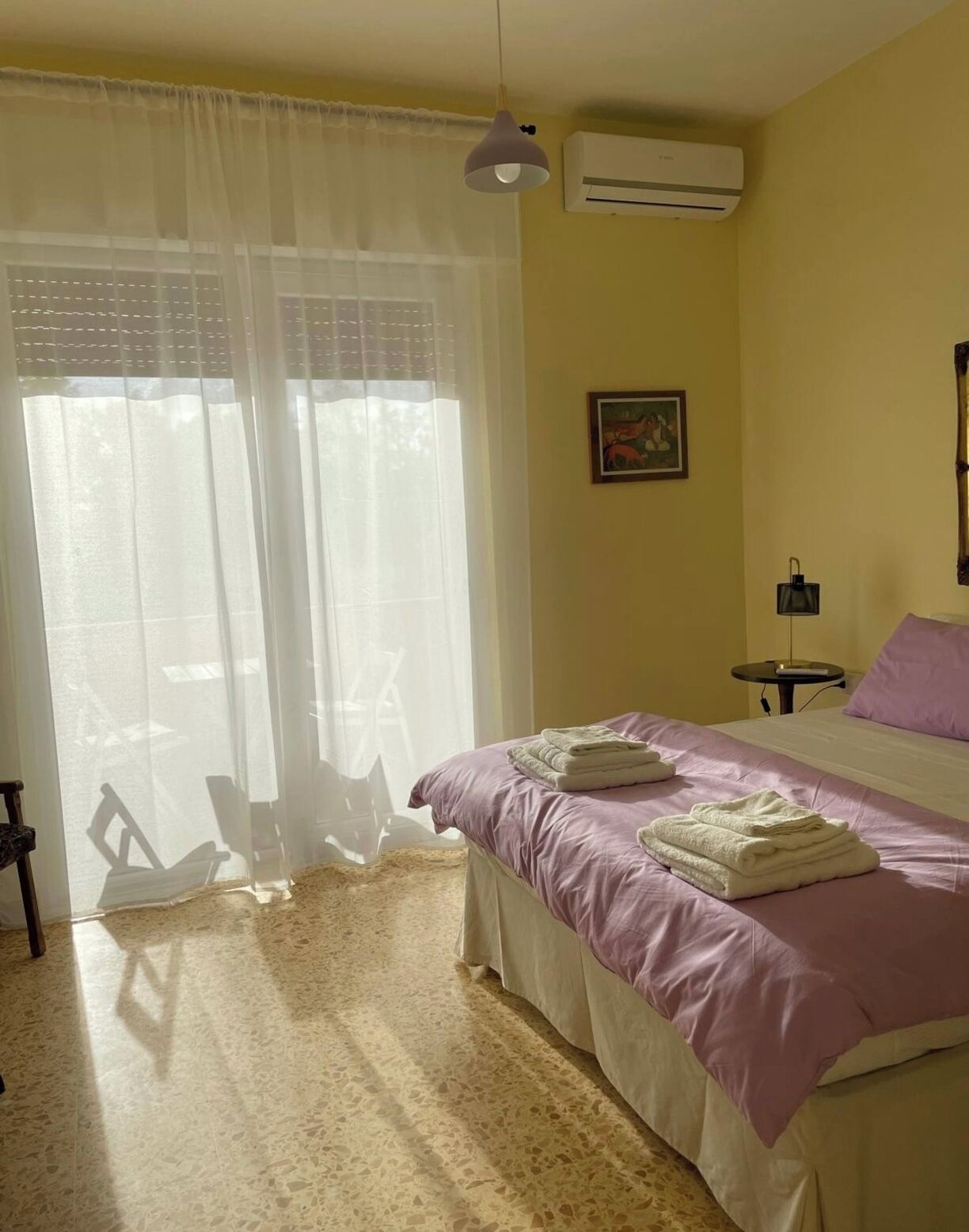 A bedroom with a bed, a chair, a table, an air conditioner, and a window with sheer curtains.