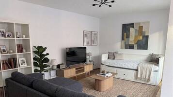 Ein modernes Wohnzimmer mit einer Couch, einem Fernseher, einem Bücherregal und einem Bett, versehen mit einem Wandkunstwerk im geometrischen Stil.