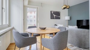 A modern living room with a round wooden table, grey upholstered chairs, a grey sofa, a flat-screen TV, and a pendant light.