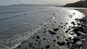 A beach with waves, rocks, and a distant view of hills. - Nestled in serene countryside, this secluded cottage boasts cosy interiors. (Llangefni)