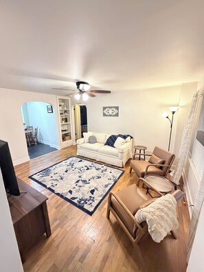 Ein Wohnzimmer mit einem weißen Sofa, einem Couchtisch, einem beige farbenen Sessel, einer Stehlampe und einem Deckenventilator.