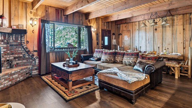 A rustic interior with a brick fireplace, wooden ceiling beams, a leather sofa, and a dining table. - Vaulted Wood Ceilings + Pine-Shaded Decks | Slopes + Trails | Pinewood Perch (Big Bear)