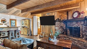 A rustic interior with a wooden ceiling, a fireplace, a flat-screen TV, and a cozy seating area with a patterned rug. - Vaulted Wood Ceilings + Pine-Shaded Decks | Slopes + Trails | Pinewood Perch (Big Bear)