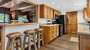 A kitchen with wooden cabinets, a refrigerator, a microwave, and a bar area with stools. - Vaulted Wood Ceilings + Pine-Shaded Decks | Slopes + Trails | Pinewood Perch (Big Bear)