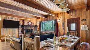 A wooden interior with a fireplace, seating area, and a dining table set for a meal. - Vaulted Wood Ceilings + Pine-Shaded Decks | Slopes + Trails | Pinewood Perch (Big Bear)