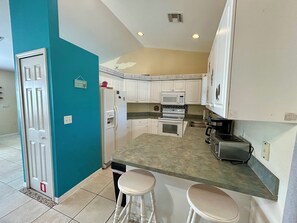 A compact kitchen with white cabinets, a built-in microwave, a stove, and a toaster. There are two white stools and a small trash bin. - 3-bedroom pool house in SW Cape Coral - pet friendly with fenced backyard (Cape Coral)
