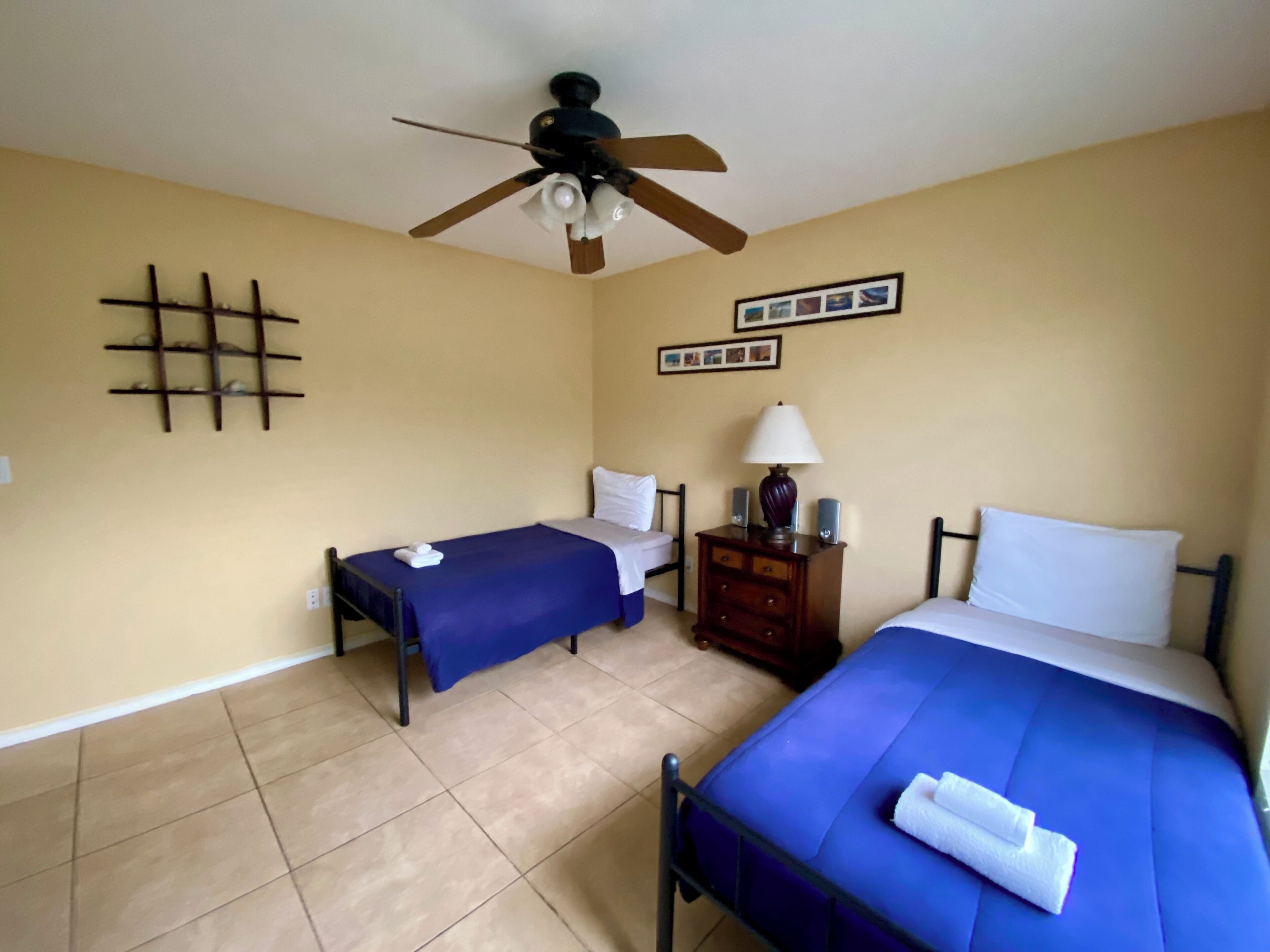 A room with two beds, a ceiling fan, a small table with a lamp, and a wall-mounted shelf.
