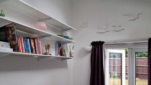Una sala de estar con un sofá gris, una estantería con libros y objetos decorativos, una ventana con cortinas y vista al jardín trasero.