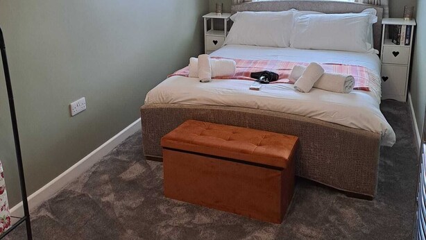 Un dormitorio con una cama grande, una mesita de noche, una lámpara de noche, un adorno redondo en la pared, una ventana con cortinas y suelo alfombrado.