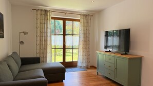 Una sala de estar con un sofá, un televisor sobre un mueble, una lámpara de pie y vistas a través de una puerta.