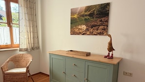 Una habitación con piso de madera, una silla de mimbre, un mueble color turquesa con cajones, un estante de madera y un cuadro colgado en la pared.