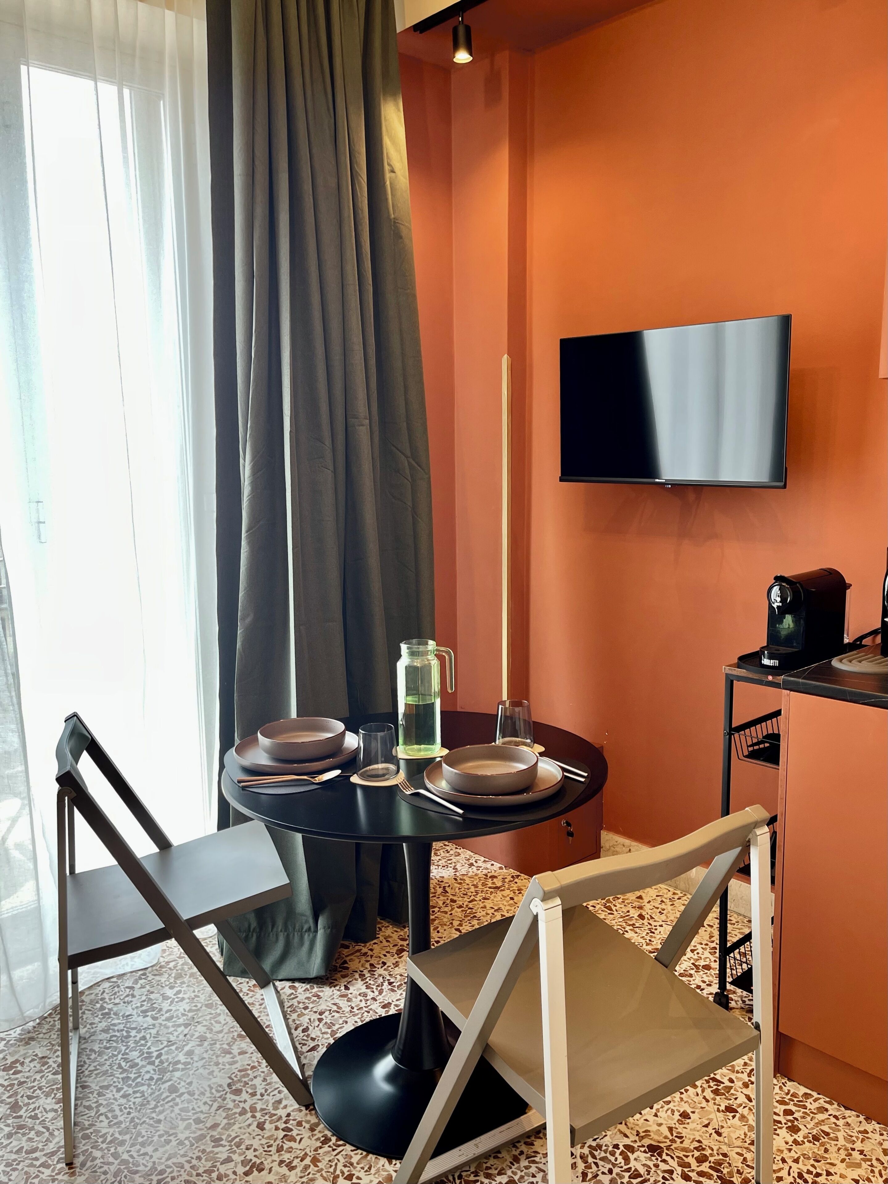A dining area with a table set for two, a television mounted on the wall, and a coffee machine on a small table.