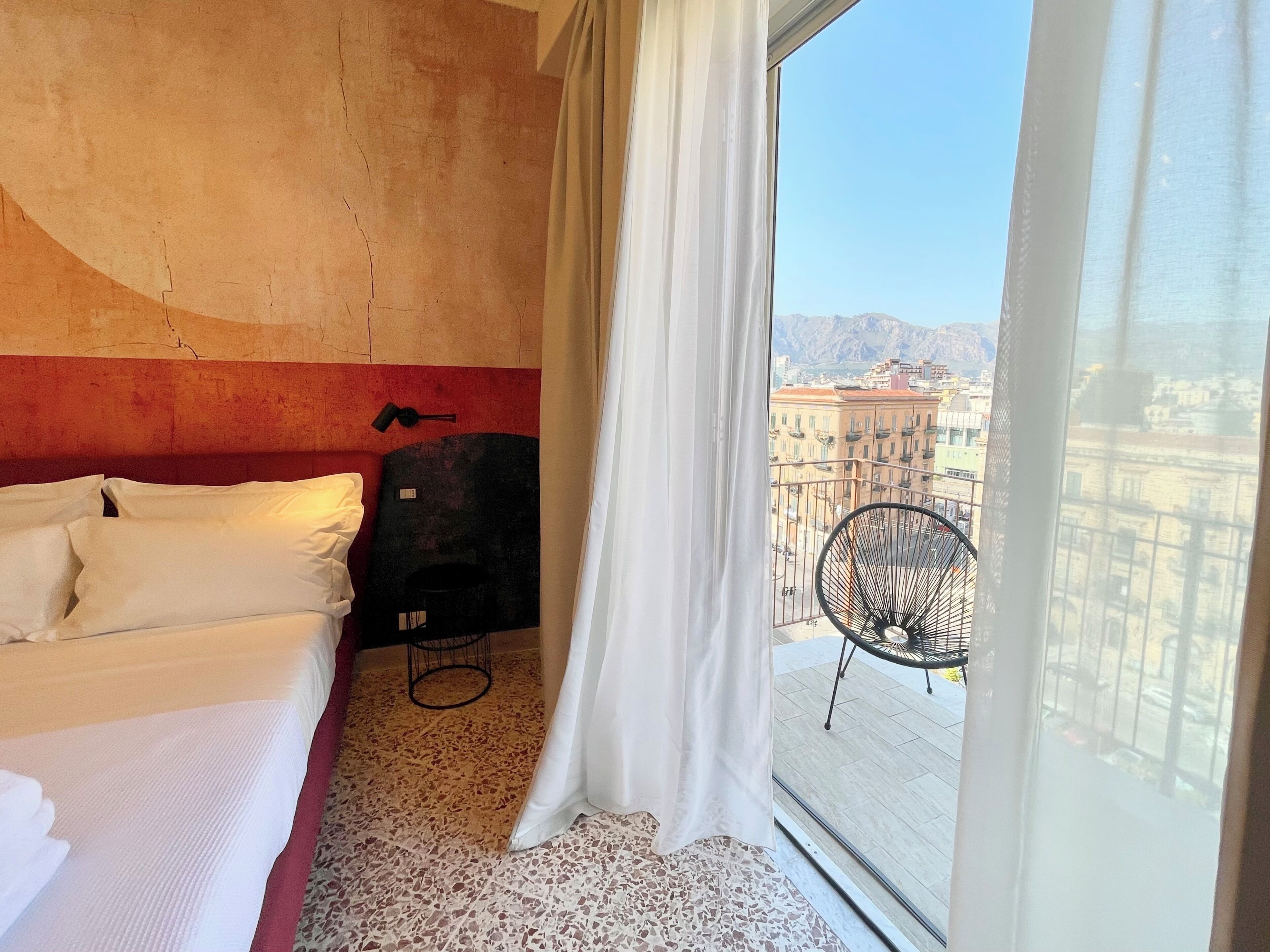 Ein Hotelzimmer mit Bett, einem Sessel und Blick auf die Stadt durch ein großes Fenster.
