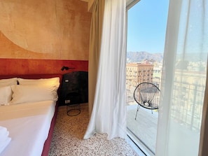 Ein Hotelzimmer mit Bett, einem Sessel und Blick auf die Stadt durch ein großes Fenster.