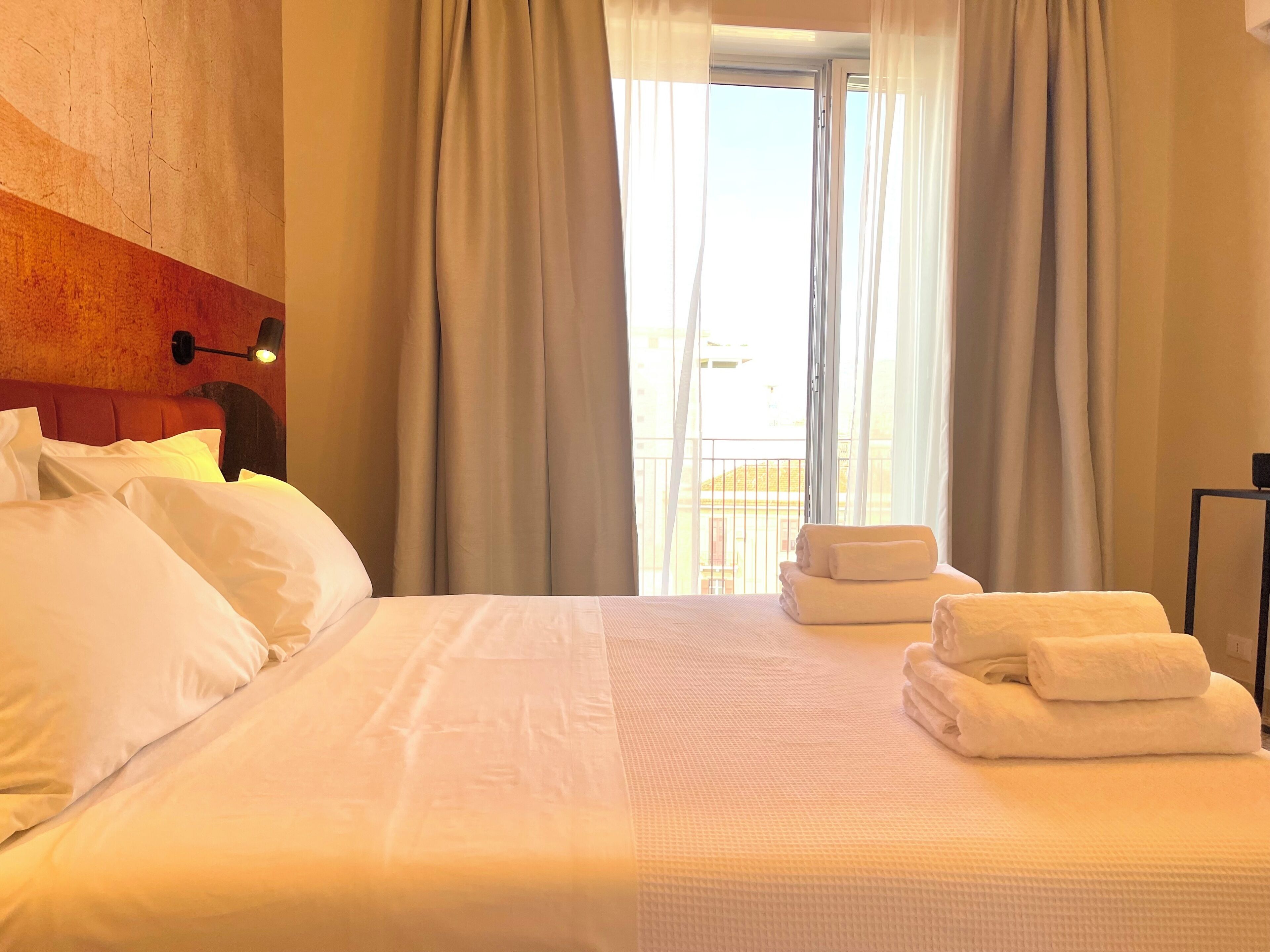 Habitación de hotel con cama, almohadas, cabecero, lámpara de pared, ventana con cortinas y balcón con barandilla.