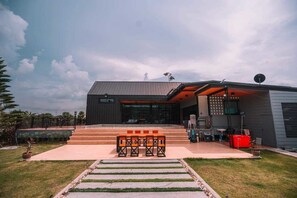 A modern building with a flat roof, large windows, and a patio area with outdoor seating. - KN Pool Villa Saraburi (Khok Yae)