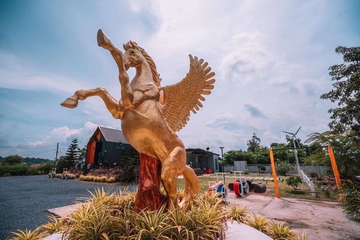 Une statue équestre dorée, représentant un cheval ailé, se trouve dans un espace paysager, avec un bâtiment en arrière-plan.
