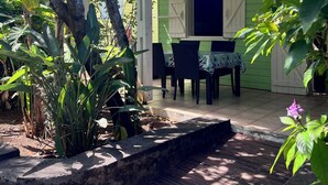 Un patio con mesa y sillas, rodeado de abundante follaje y una casa de fachada verde.
