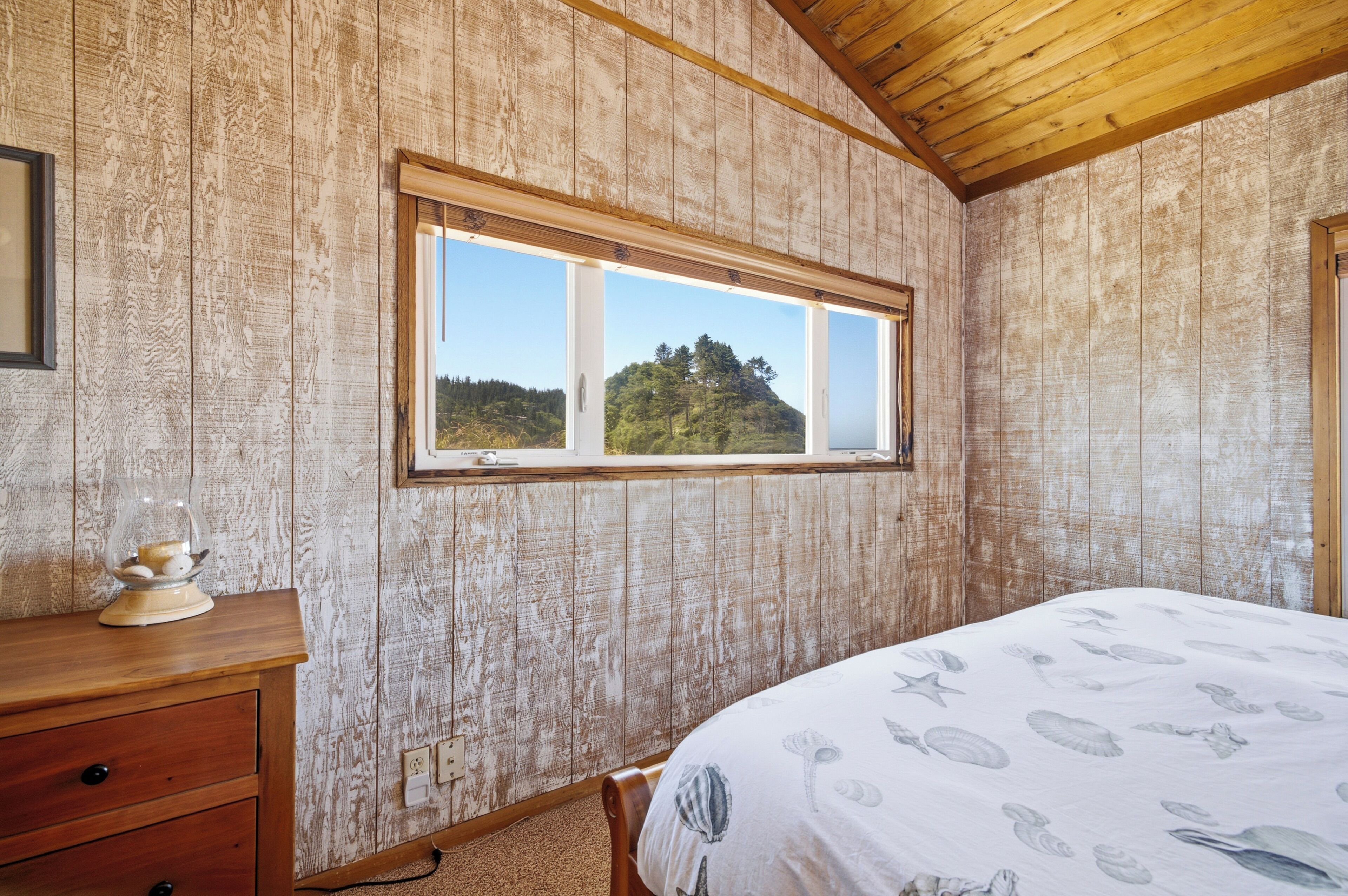 Une chambre avec un plafond en bois, une fenêtre donnant sur une colline, un lit orné d’un motif de coquillage et une commode en bois.
