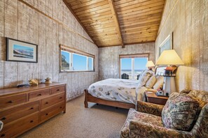 Quarto com teto de madeira, cama com colcha de conchas, cômoda, duas janelas e mesa de cabeceira com abajur.