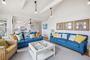 Ein Wohnzimmer mit blauen Sofas, einem weißen Couchtisch, Korbstühlen und einer Küche im Hintergrund.