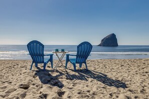 Zwei blaue Stühle und ein kleiner Tisch am Sandstrand, im Hintergrund das Meer und eine große Felsformation.
