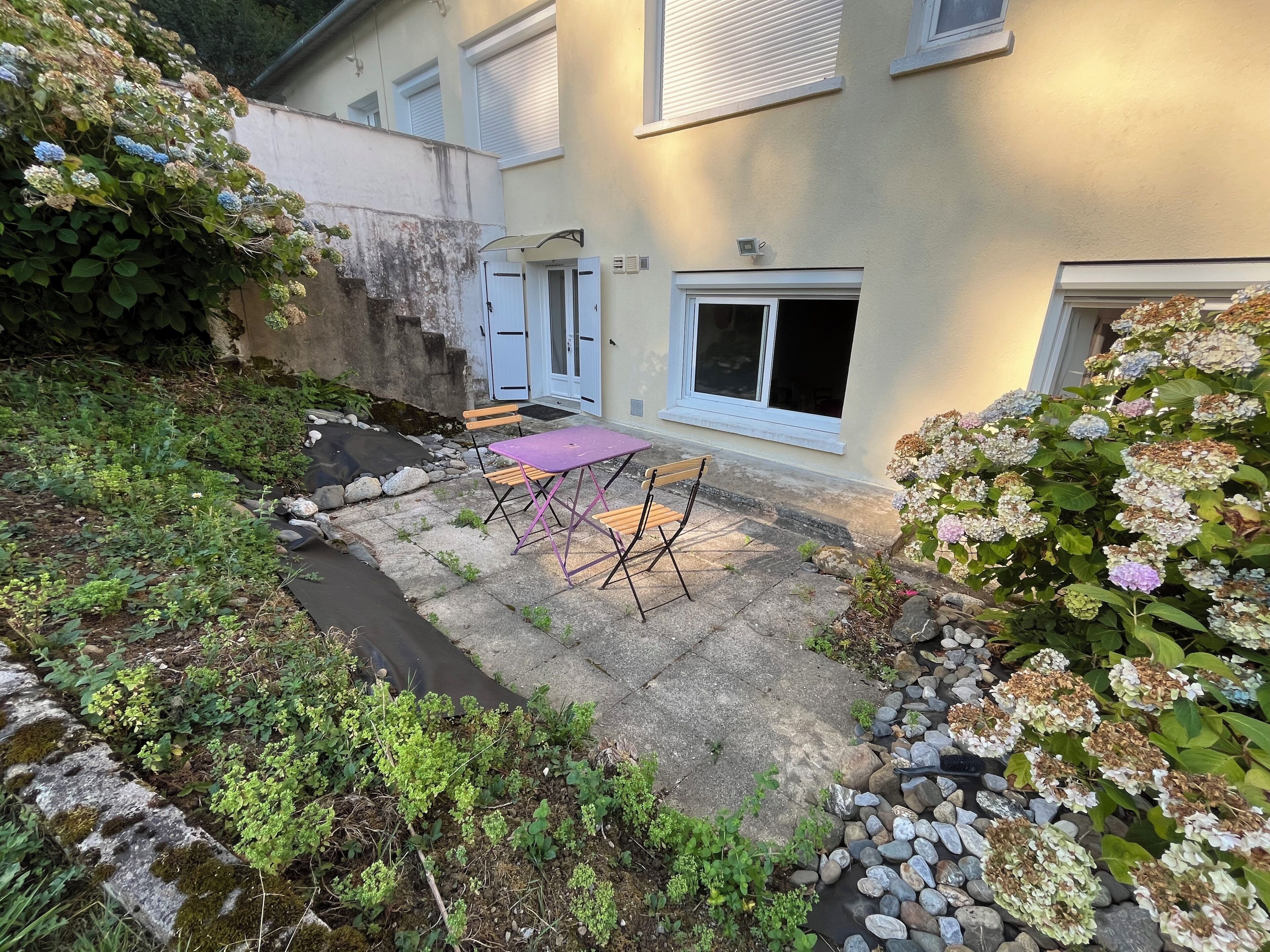 Une cour avec une terrasse en pierre, une table et des chaises violettes, le tout entouré de verdure et traversé par un petit ruisseau.