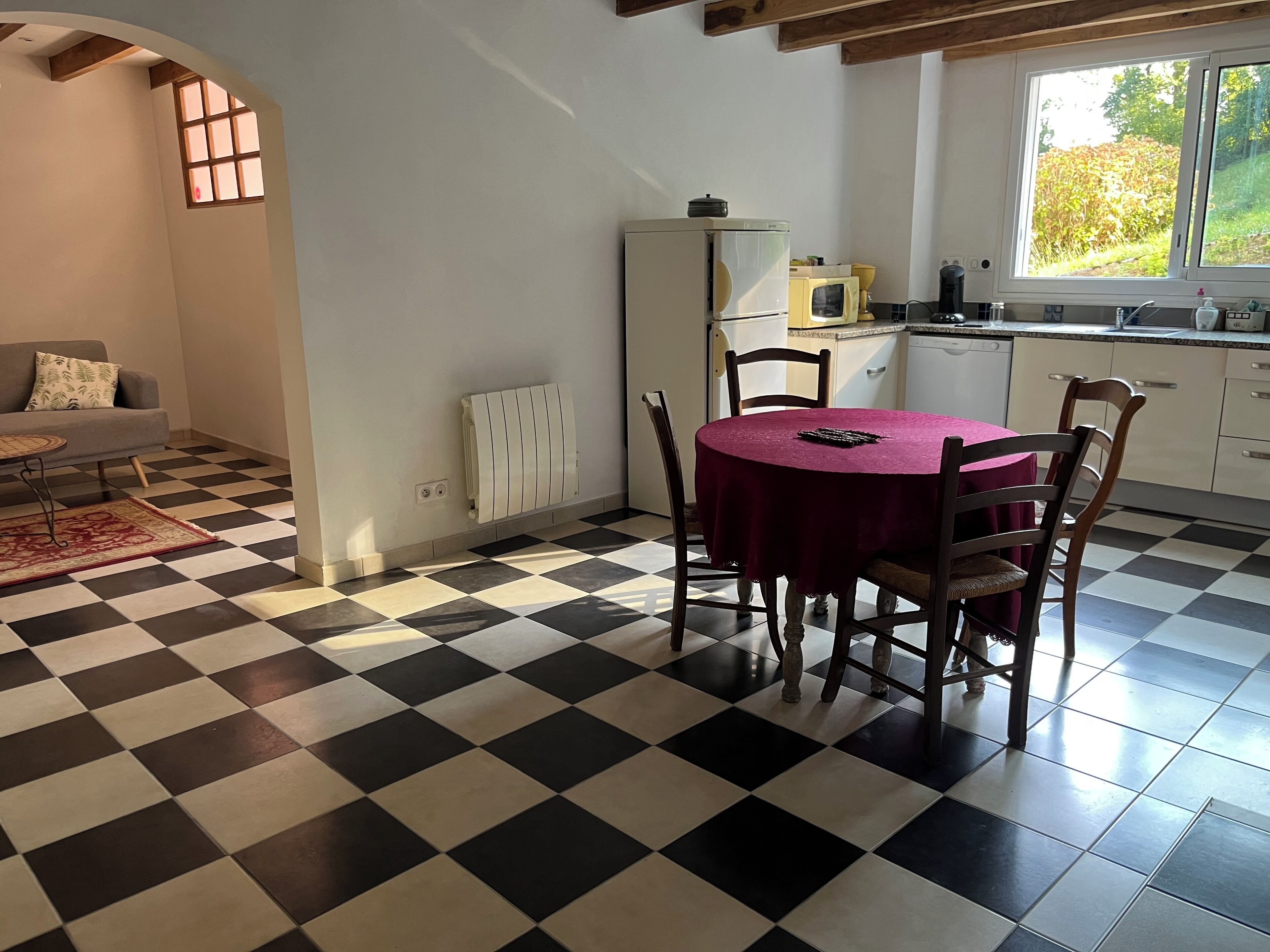 Un coin repas avec un sol à carreaux, une table ronde entourée de chaises, un radiateur et une fenêtre donnant sur un paysage verdoyant.