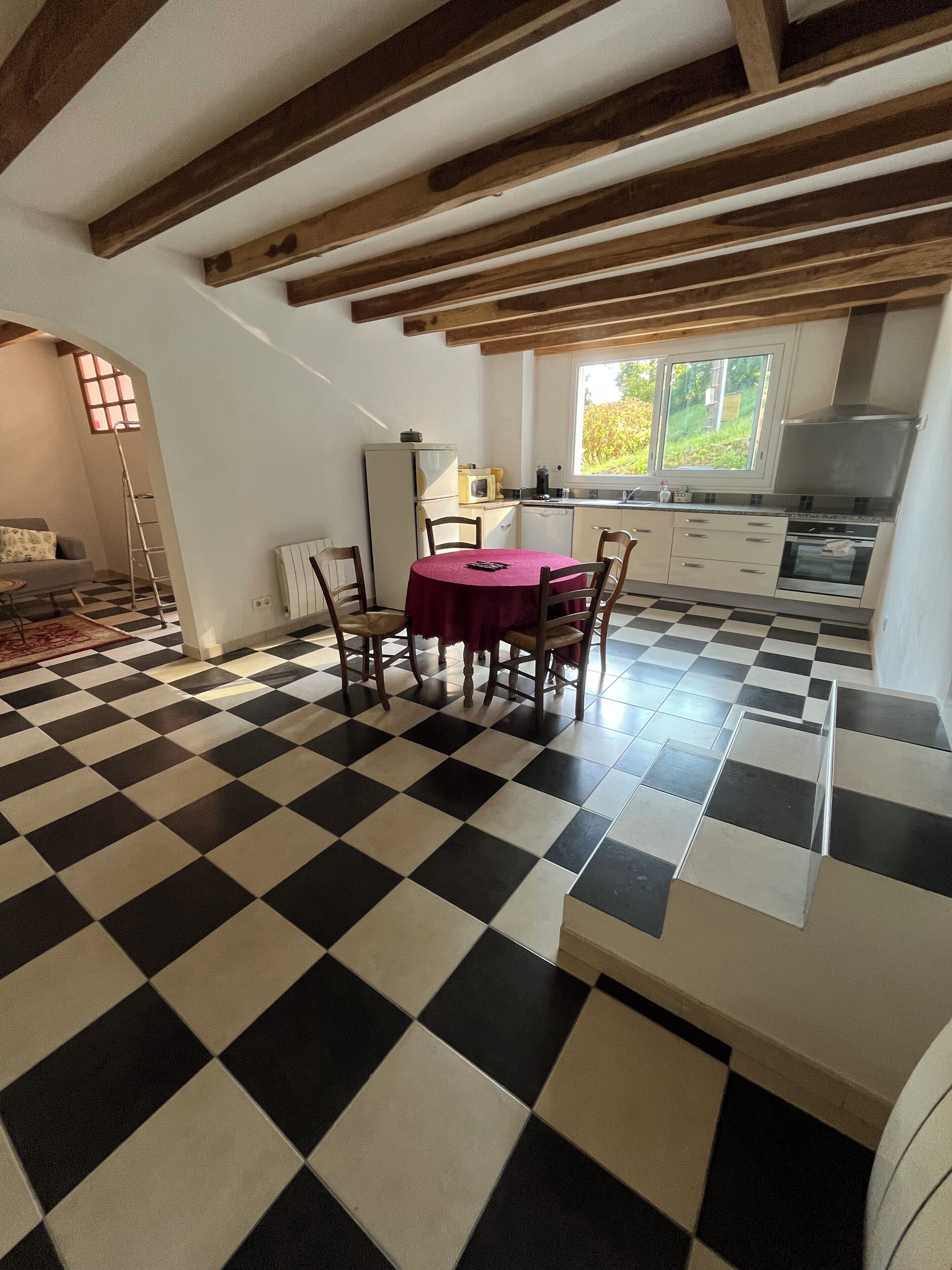 Une pièce dotée d’un sol à carreaux, d’une table à manger avec des chaises, d’un coin cuisine avec des placards et des appareils électroménagers, et d’une fenêtre donnant sur un espace verdoyant.