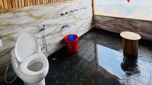 Un baño con techo de madera, pared de mármol, un inodoro, un cubo rojo y vistas al exterior por una ventana.