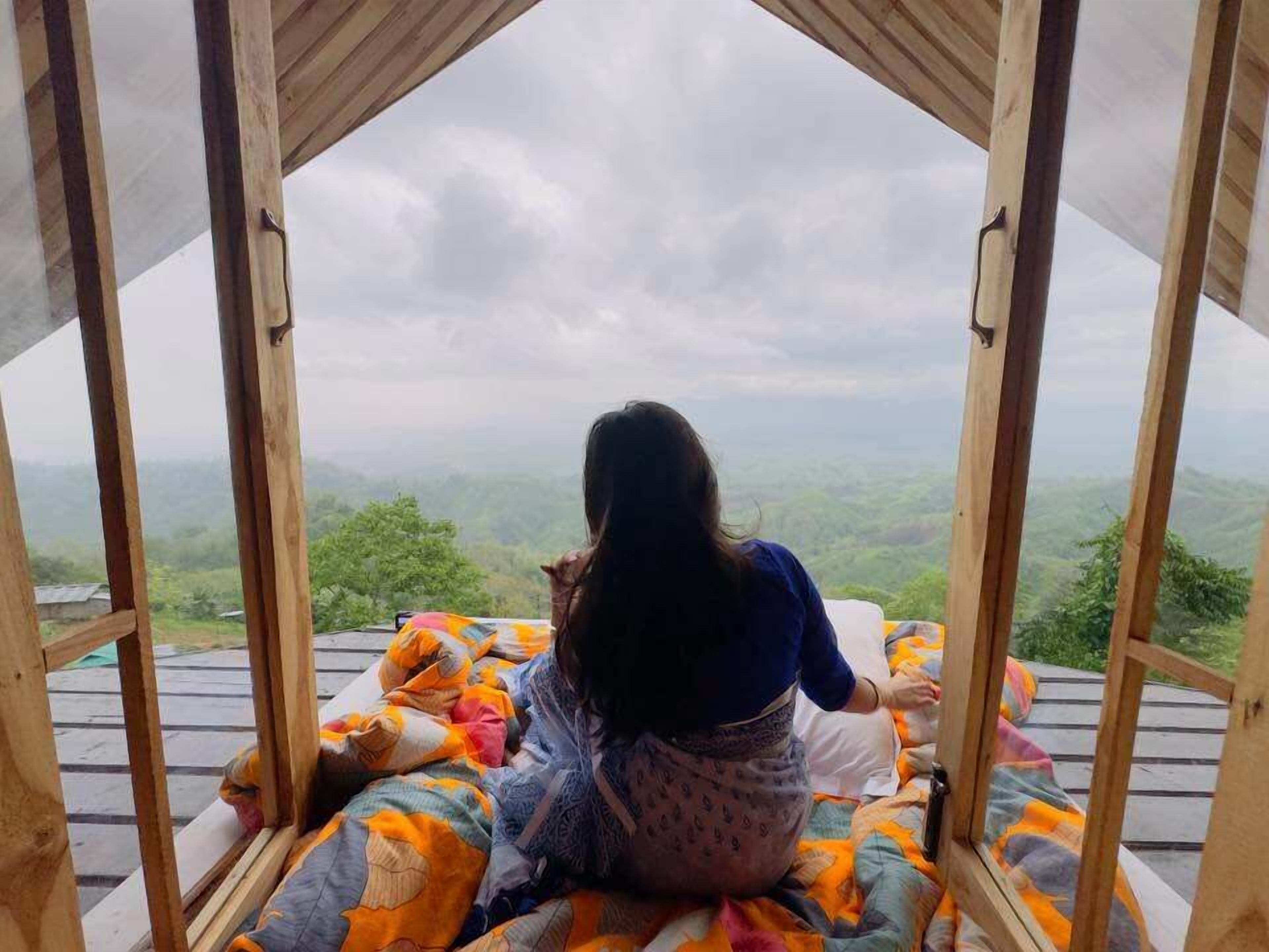A person sitting on a bed with a view of a mountainous landscape through a wooden structure.