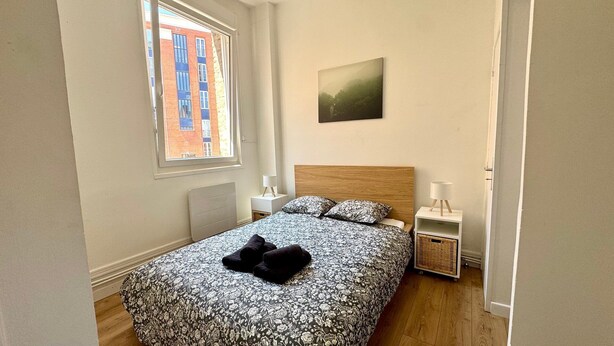 A bedroom with a bed, a nightstand, a lamp, a picture on the wall, and a window with a view of buildings. - The Baudimont First (Arras)